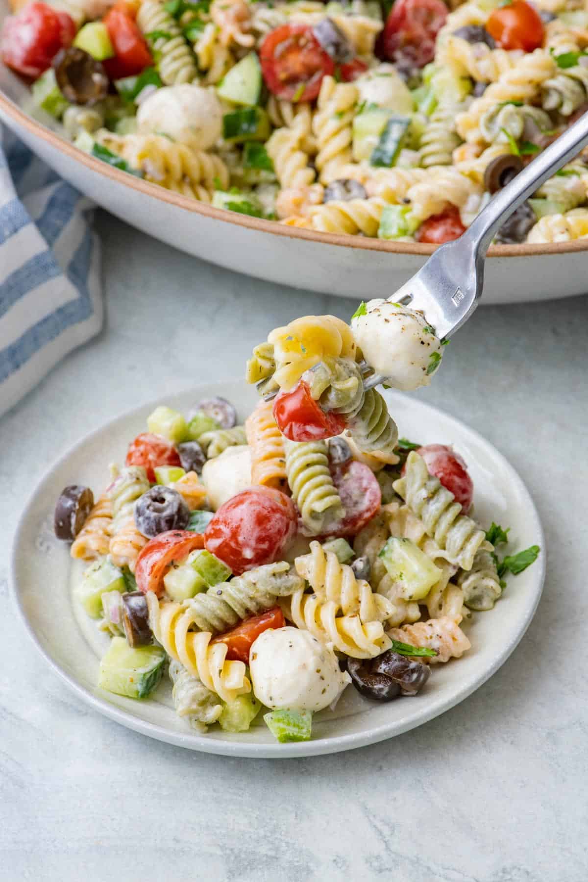 A serving of Italian pasta salad on a small plate with the serving bowl nearby. A fork is lifting up a bite.