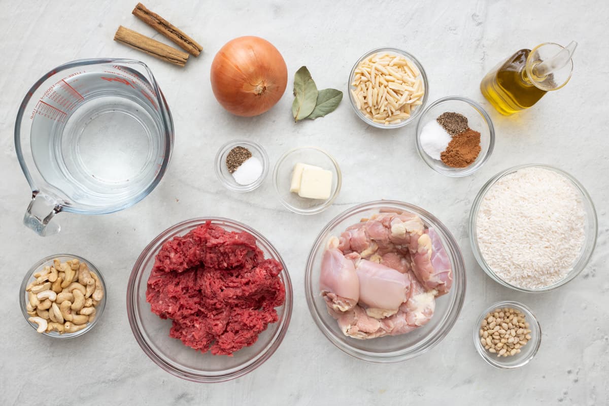 Ingredients for recipe before prepping in individual bowls: water, nuts, onion, salt and pepper, butter, bay leaves, slivered almonds, spices, oil, rice, pinenuts, bonesless chicken thighs, and ground beef.