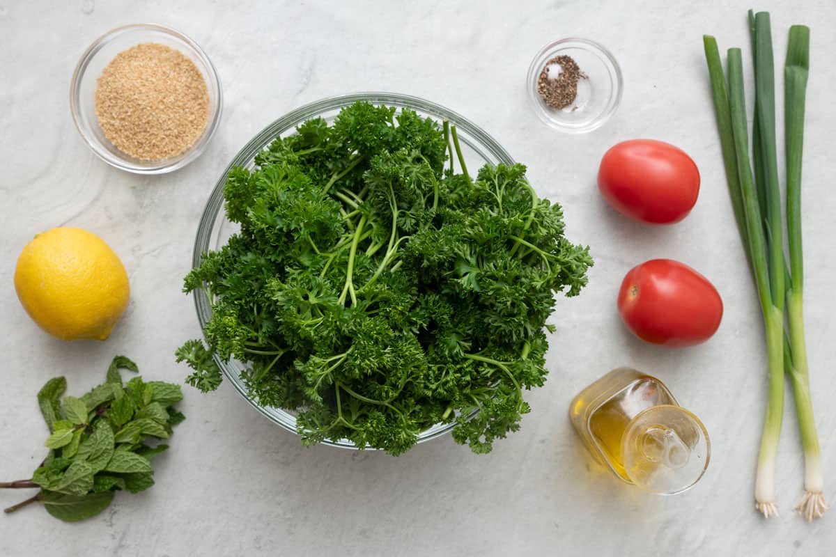 Ingredients for recipe before prepping: fine bulgur, lemon, mint, curly parsley, salt and pepper, roma tomatoes, oil, and green onions.