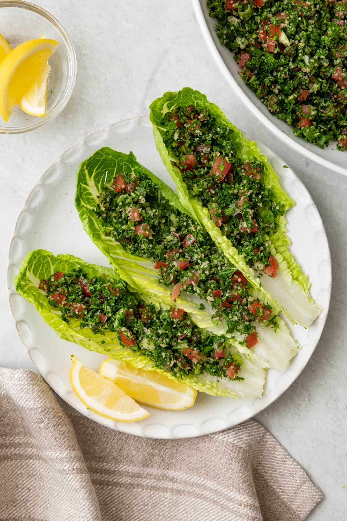 3 romaine lettuce cups filled with tabbouleh salad on a serving plate with lemon wedges and the bowl of salad nearby.
