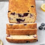 Loaf of lemon blueberry bread with 3 thick slices cut away from loaf on a parchment line wire rack with fresh lemon slices and blueberries around.