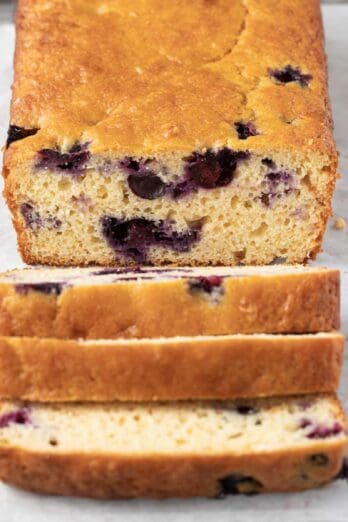 Loaf of lemon blueberry bread with 3 thick slices cut away from loaf on a parchment line wire rack with fresh lemon slices and blueberries around.