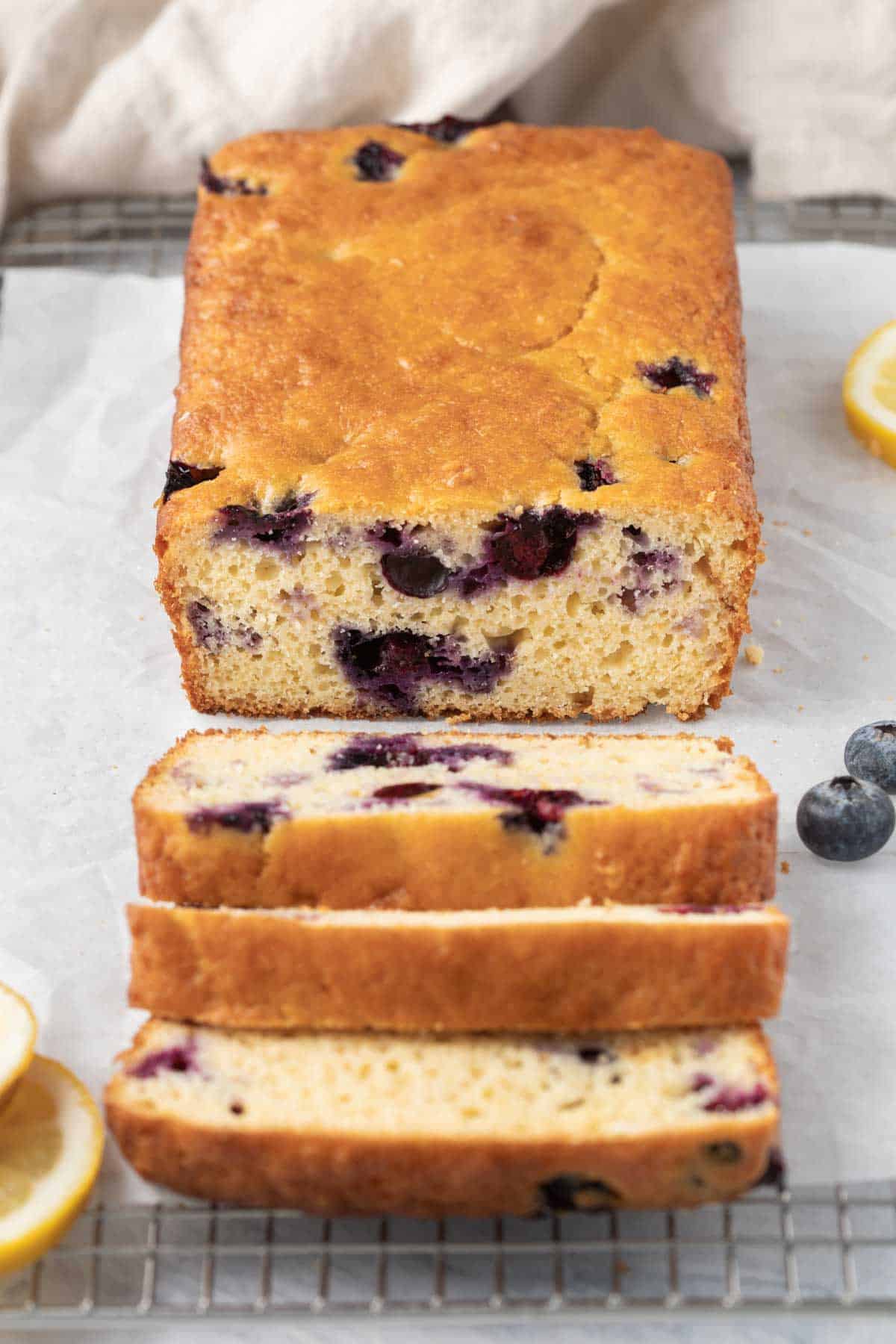Lemon blueberry loaf cut halfway on wire wrack with fresh lemons nearby.