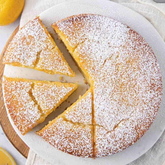 Whole lemon cake sprinkled with powdered sugar with three slices cut from one side, two slices are pulled away slightly and the third is still attached to the cake. Cake plate is sitting on a round wooden board with a towel over it, with lemon wedges around.