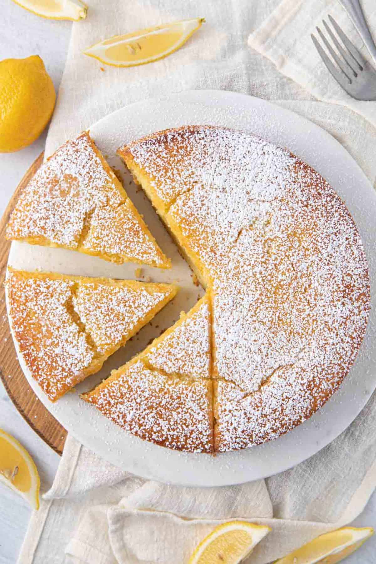 Whole lemon cake sprinkled with powdered sugar with three slices cut from one side, two slices are pulled away slightly and the third is still attached to the cake. Cake plate is sitting on a round wooden board with a towel over it, with lemon wedges around.