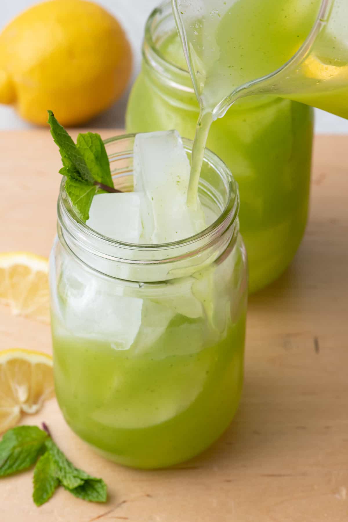 Mint lemonade being poured into an ice filled mason jar with a full glass behind it.