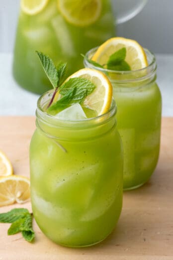 2 glasses of freshly made mint lemonade with a bright green color garnished with extra fresh lemon slices and mint and the pitcher sitting behind them.