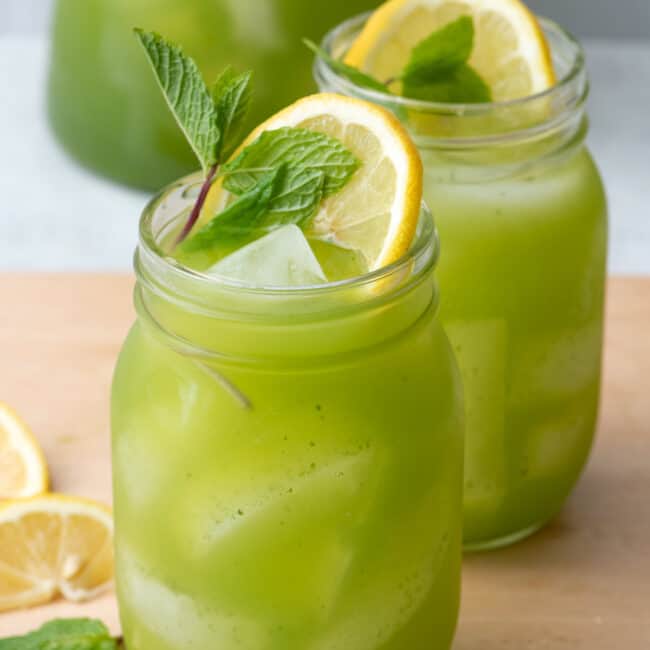 2 glasses of freshly made mint lemonade with a bright green color garnished with extra fresh lemon slices and mint and the pitcher sitting behind them.