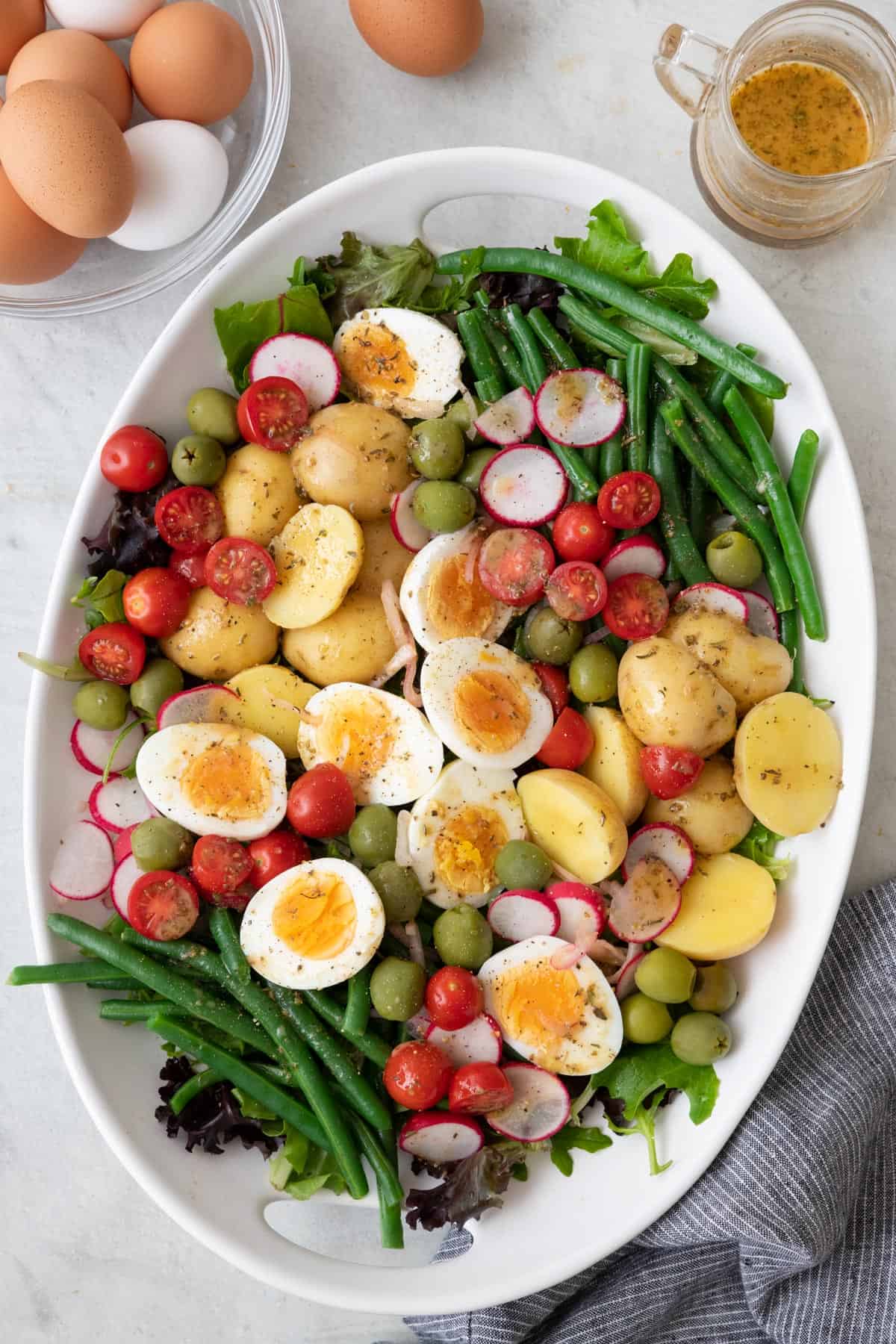 Nicoise salad ingredients layered on a large oval platter with a bowl of fresh eggs and dressing nearby.