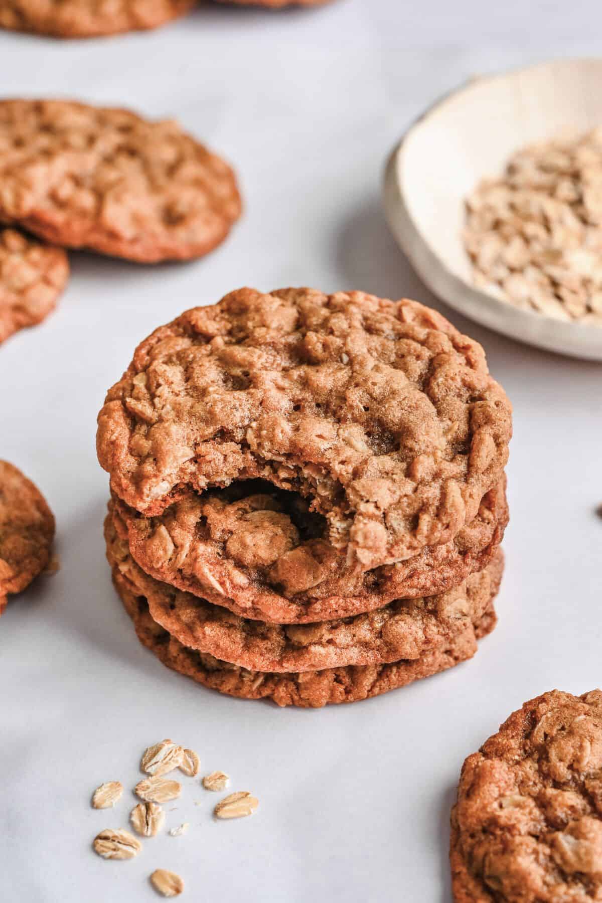 Stack of oatmeal cookies with a bite taken out of the top cookie. A small bowl of oats and more cookies around.