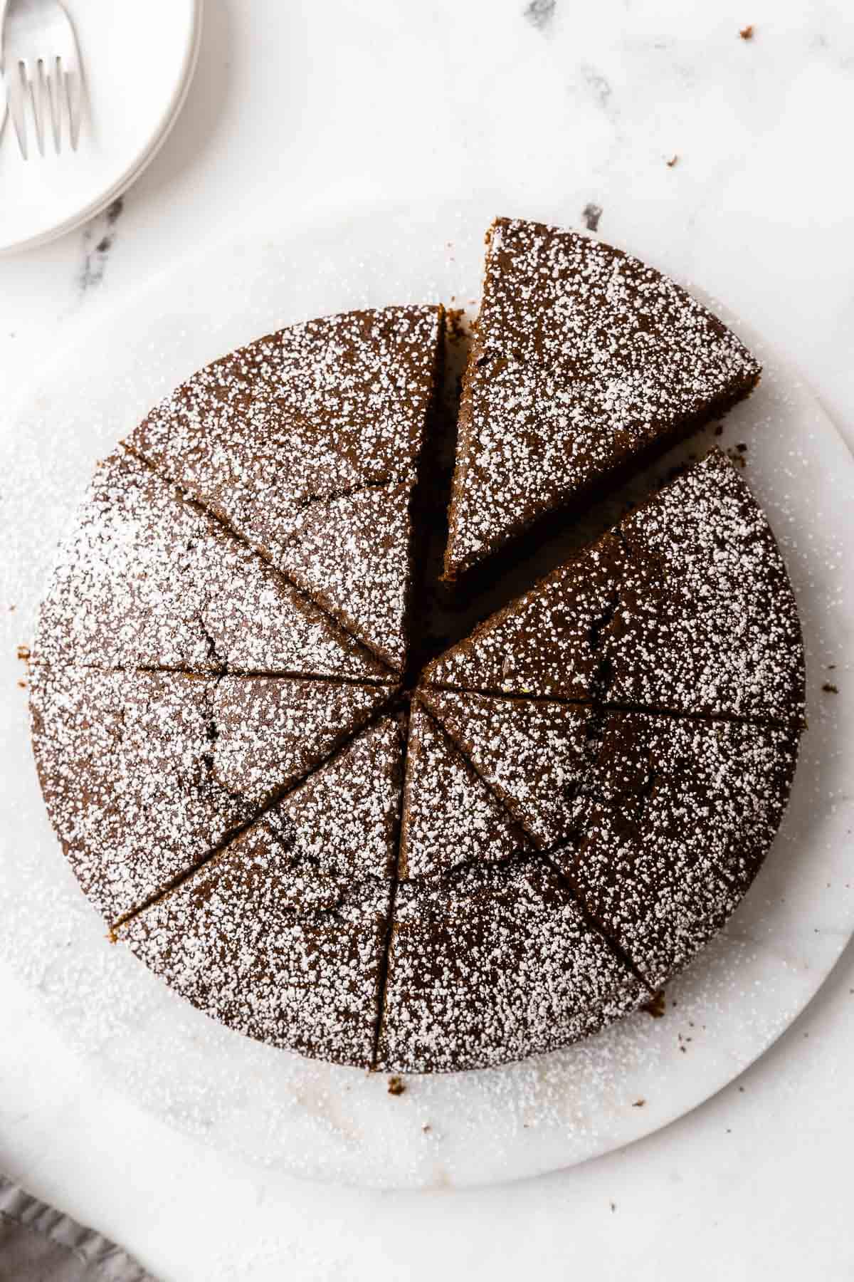 Overhead shot of chocolate olive oil cake cut into 8 slices and sprinkled with powdered sugar, sitting on marble with white plates and forks nearby.