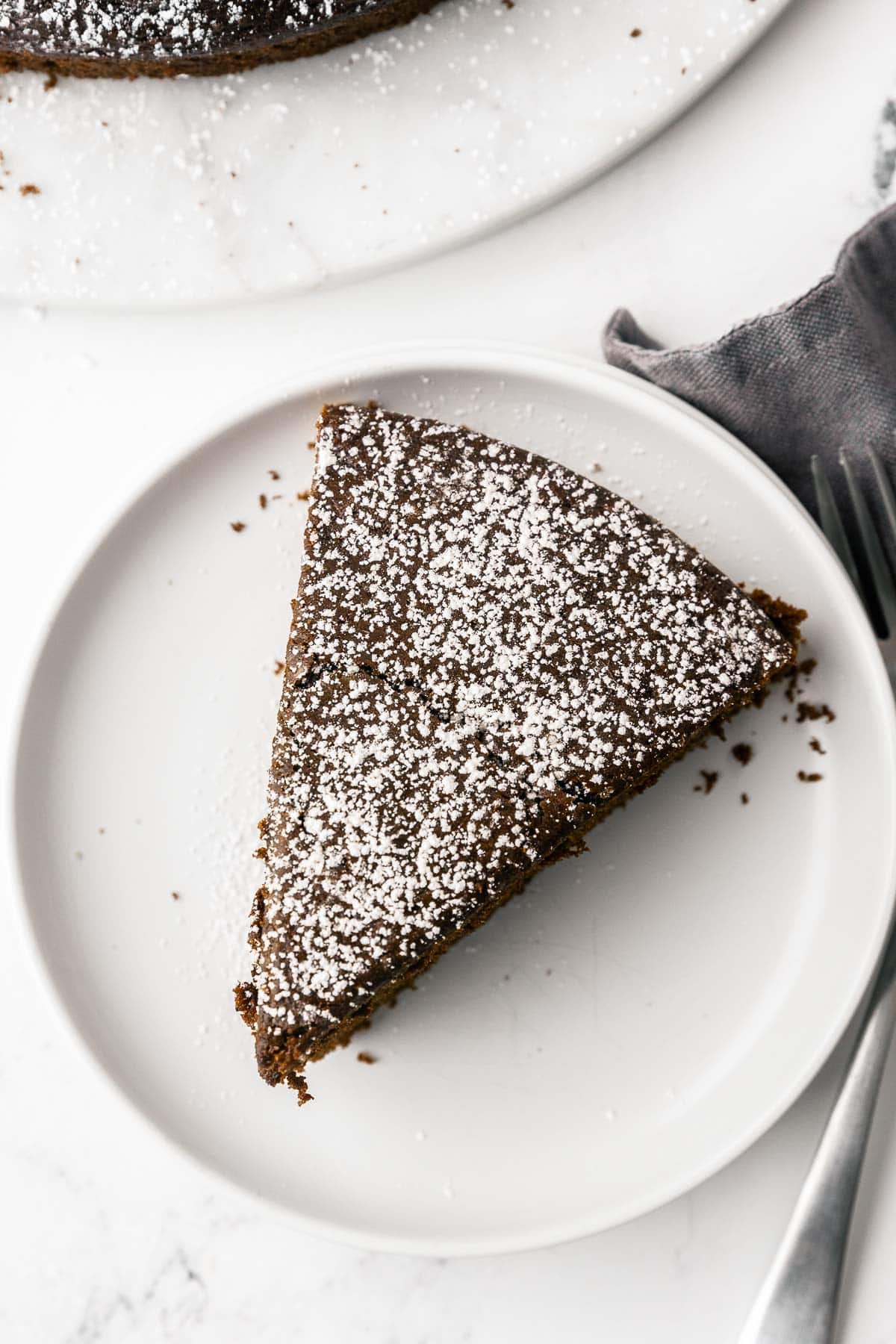 Overhead shot of a slice of chocolate olive oil cake with powdered sugar on a small white plate, forks and napkin nearby.