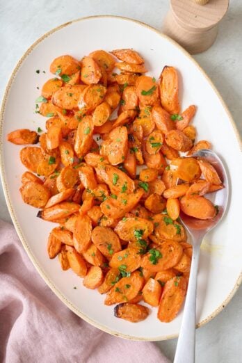 Oven roasted carrot slices on a large oval platter, garnished with fresh chopped parsley and a serving spoon dipped in carrots.