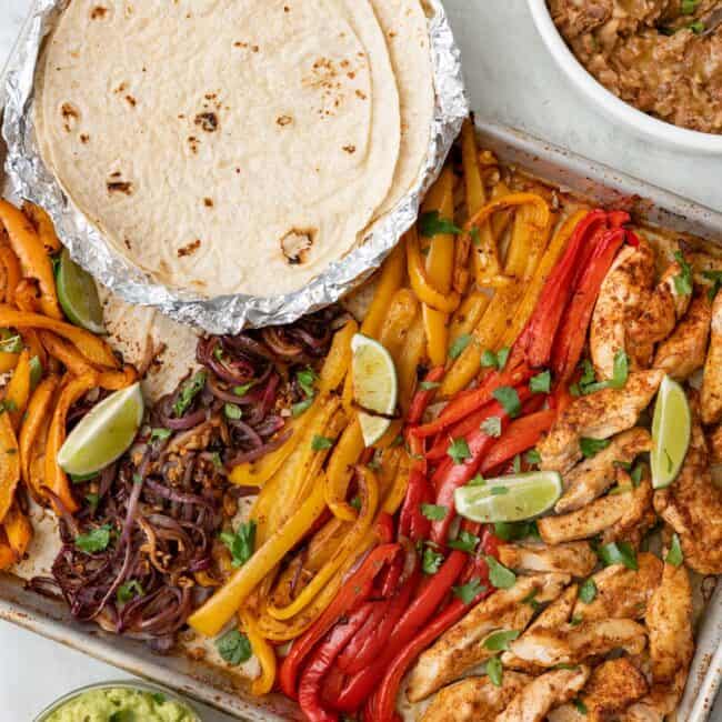 Sheet pan chicken fajitas garnished with fresh cilantro with small dishes of guacamole, salsa, flour taco shells, refried beans and sliced avocados.