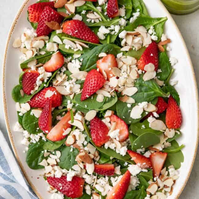 Spinach strawberry salad on a large oval platter topped with feta and sliced almonds with a basil vinaigrette nearby.