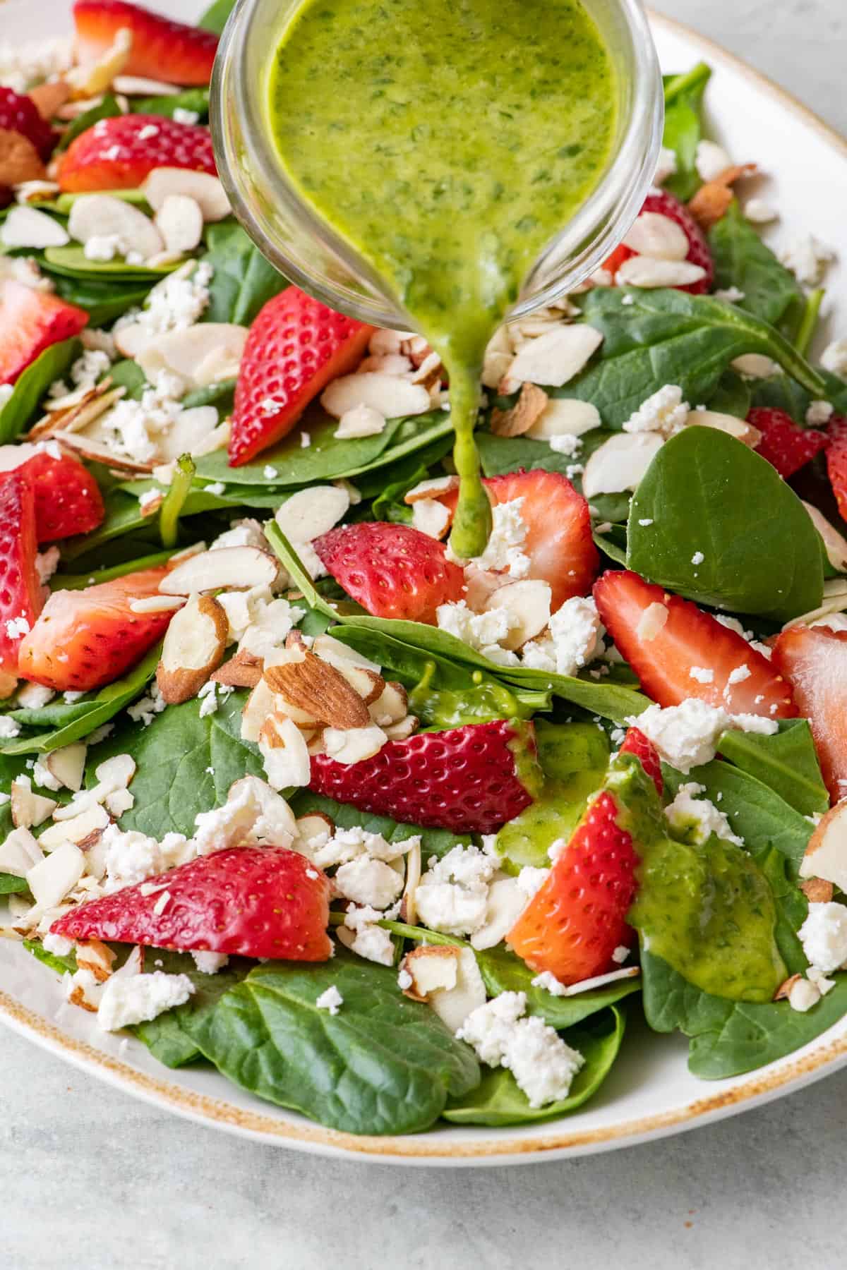 Basil vinaigrette being poured over a spinach salad with strawberries, feta, and almonds.
