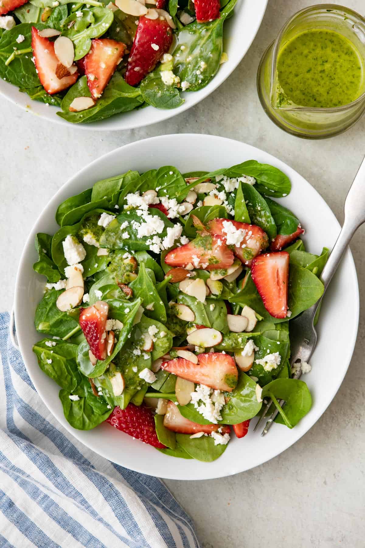 Two bowls of spinach strawberry salad with a bright green vinaigrette in a glass jar nearby.