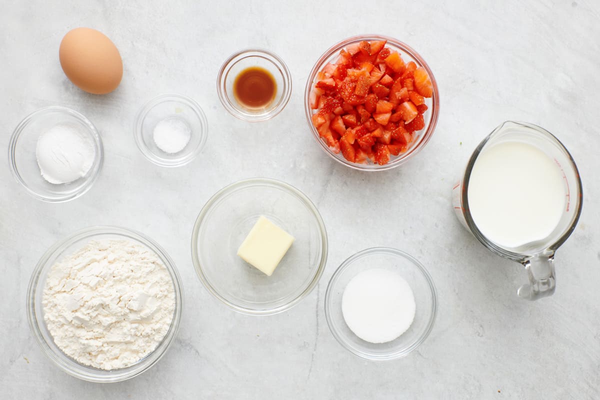 Ingredients for recipe in individual bowls: an egg, baking powder, salt, flour, butter, vanilla, diced strawberries, sugar, and milk.