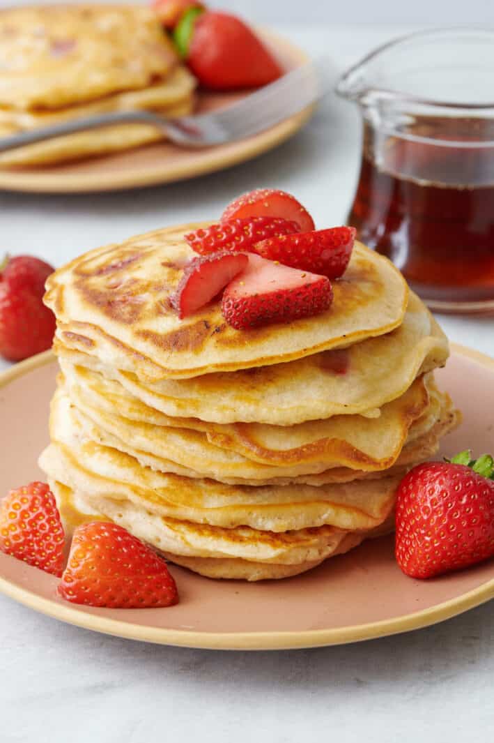 Strawberry pancakes stacked up on a plate with more fresh strawberries on top and around plate. Maple syrup and more pancakes nearby.