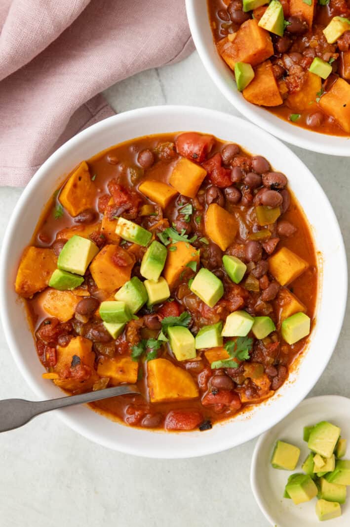 Chunky sweet potato chili in a tomato based broth with beans, topped with diced avocado with another bowl nearby.