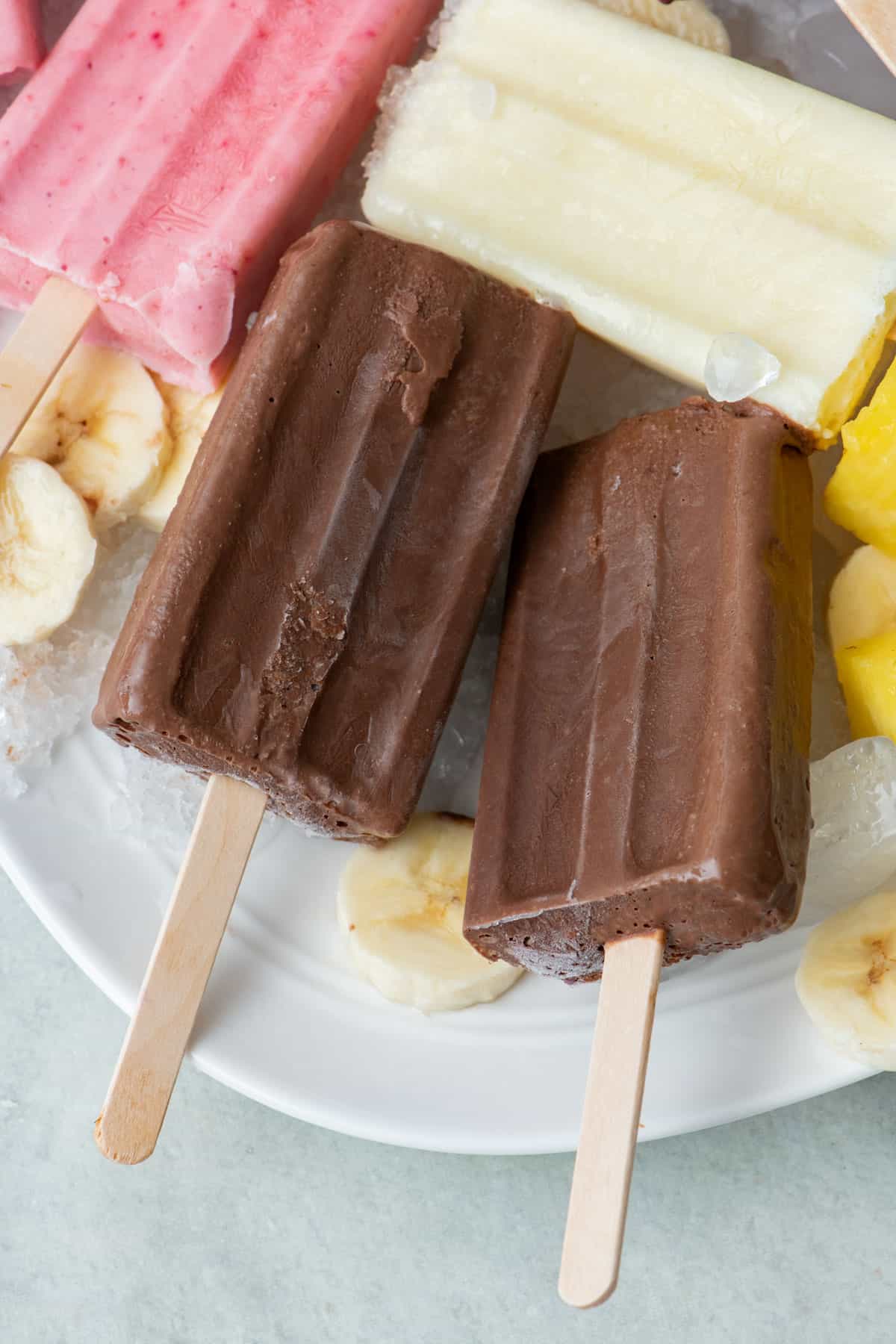 Frozen chocolate banana yogurt popsicles on a tray of ice with extra banana slices around.