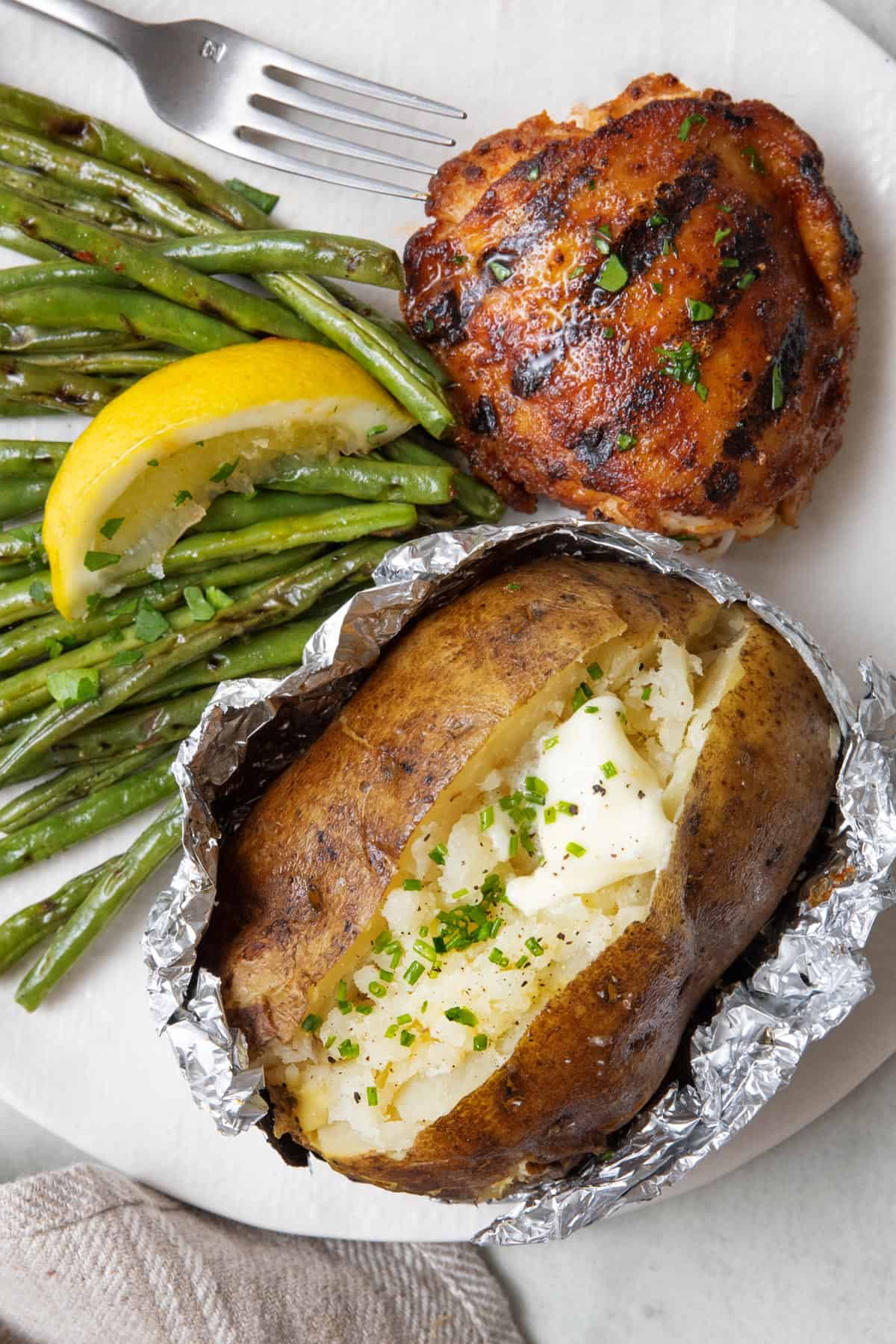 Grilled baked potato in foil split open and fluffed with butter, chives and salt plated with a grilled chicken thigh and fresh grilled string beans with a lemon wedge on top.