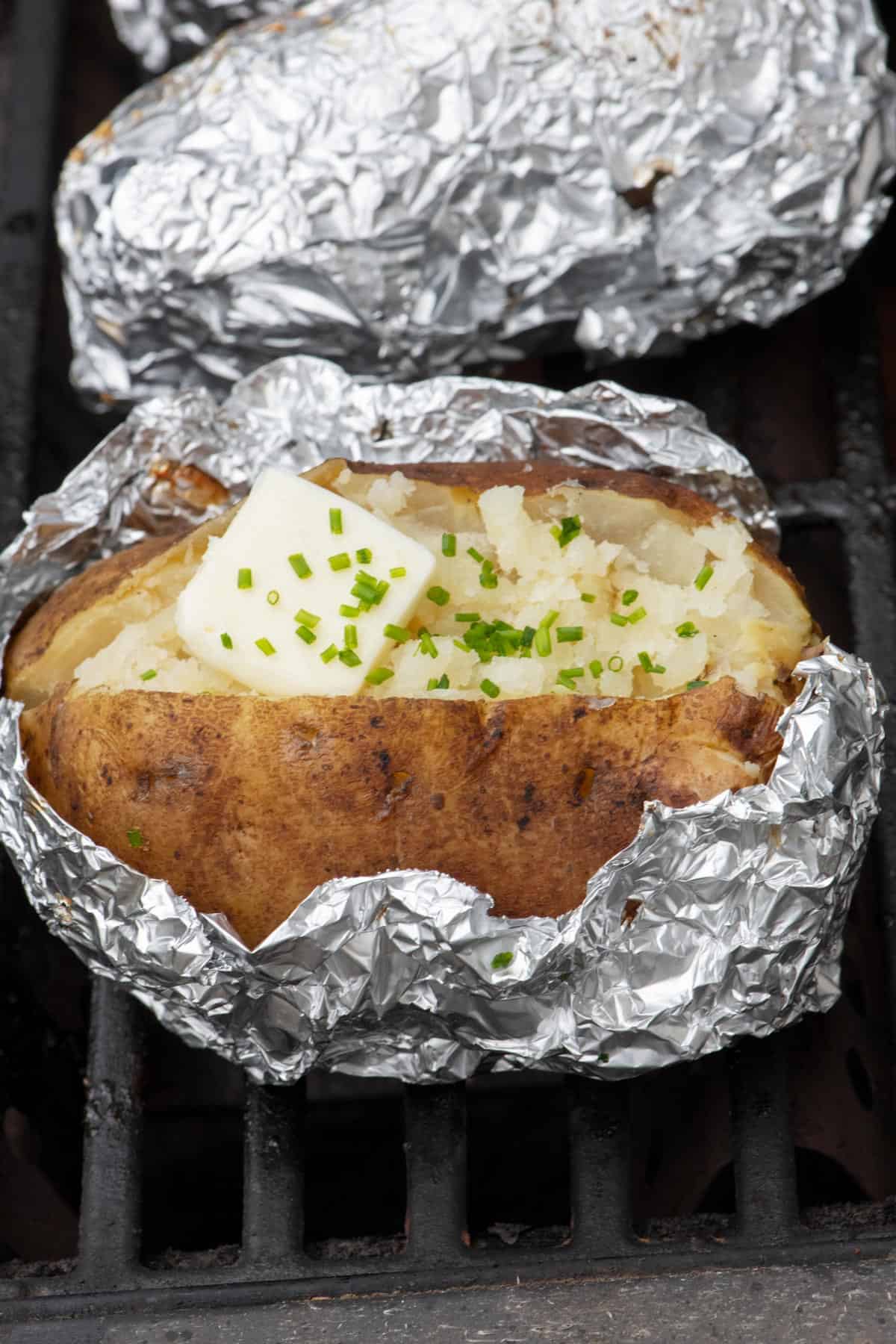 Foil wrapped baked on potatoes on the girll with one with the foil partially opened showing a split potato with butter and green onions.