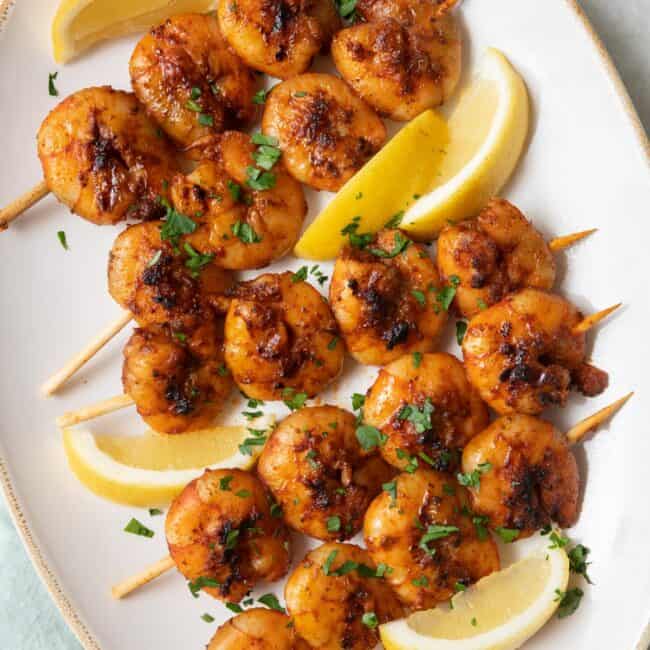Grilled BBQ shrimp skewers on a large oval platter garnished with chopped fresh parsley and lemon wedges.