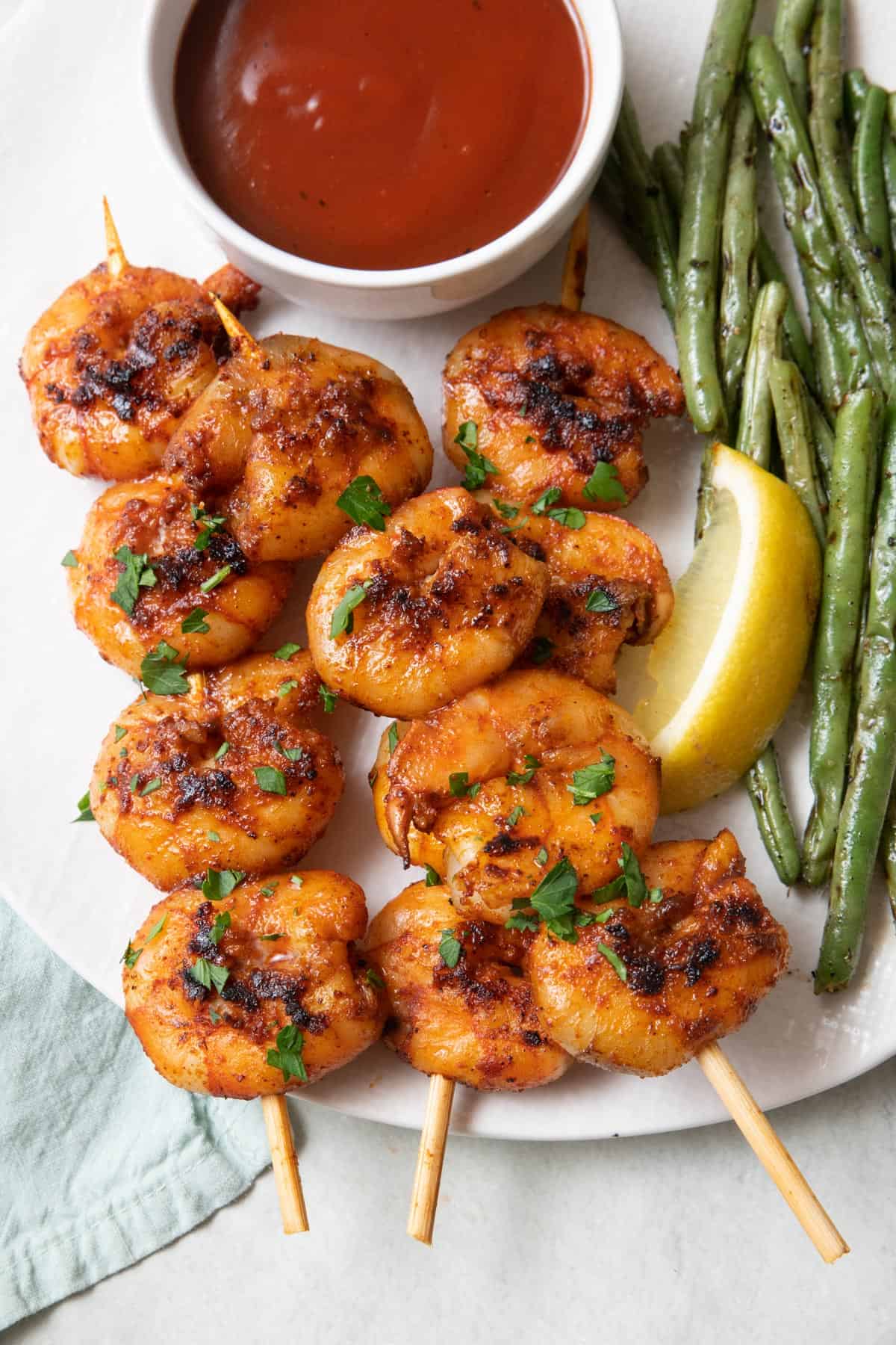 Barbeque grilled shrimp on wooden skewers on a plate with grilled green beans, a small dish of bbq sauce, and lemon wedge.