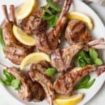 Grilled lamb chops on a round white plate garnished with mint leaves and lemon wedges.