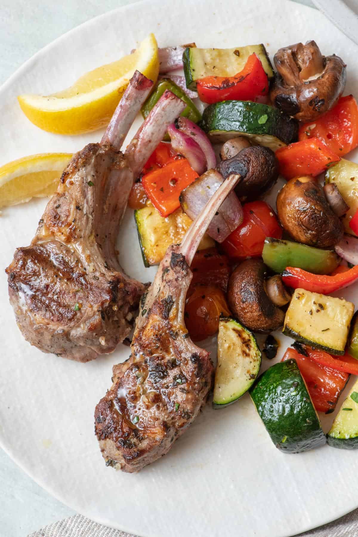 Grilled lamb chops on a serving plate with grilled veggies and lemon wedges.