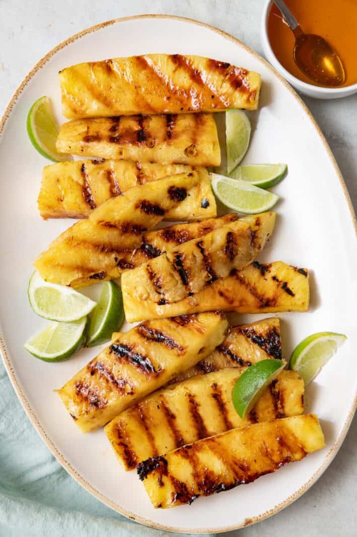 Grilled pineapple spears on a large oval platter showing garnished with fresh lime wedges with a small dish of honey nearby with a spoon dipped inside.