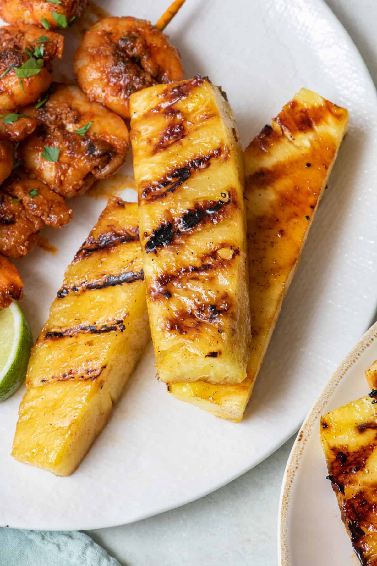Three grilled pineapple spears on a plate next to shrimp skewers.