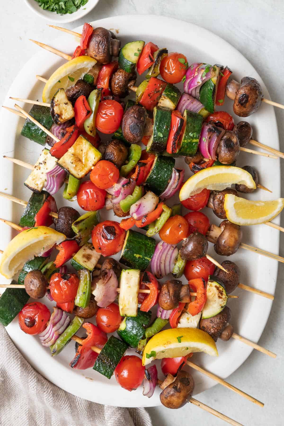 Grilled veggie skewers on a large oval platter garnished with fresh lemon wedges and chopped parsley.