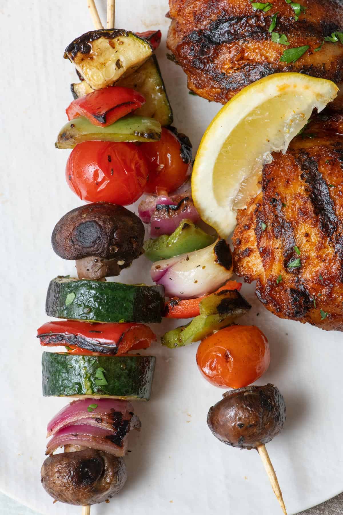 Two veggies skewers on a plate with grilled chicken and a lemon wedge.