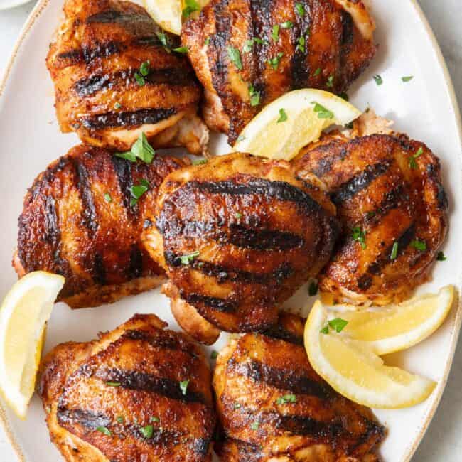 Grilled chicken thighs on an oval platter with crispy skin garnished with fresh parsley and lemon wedges.