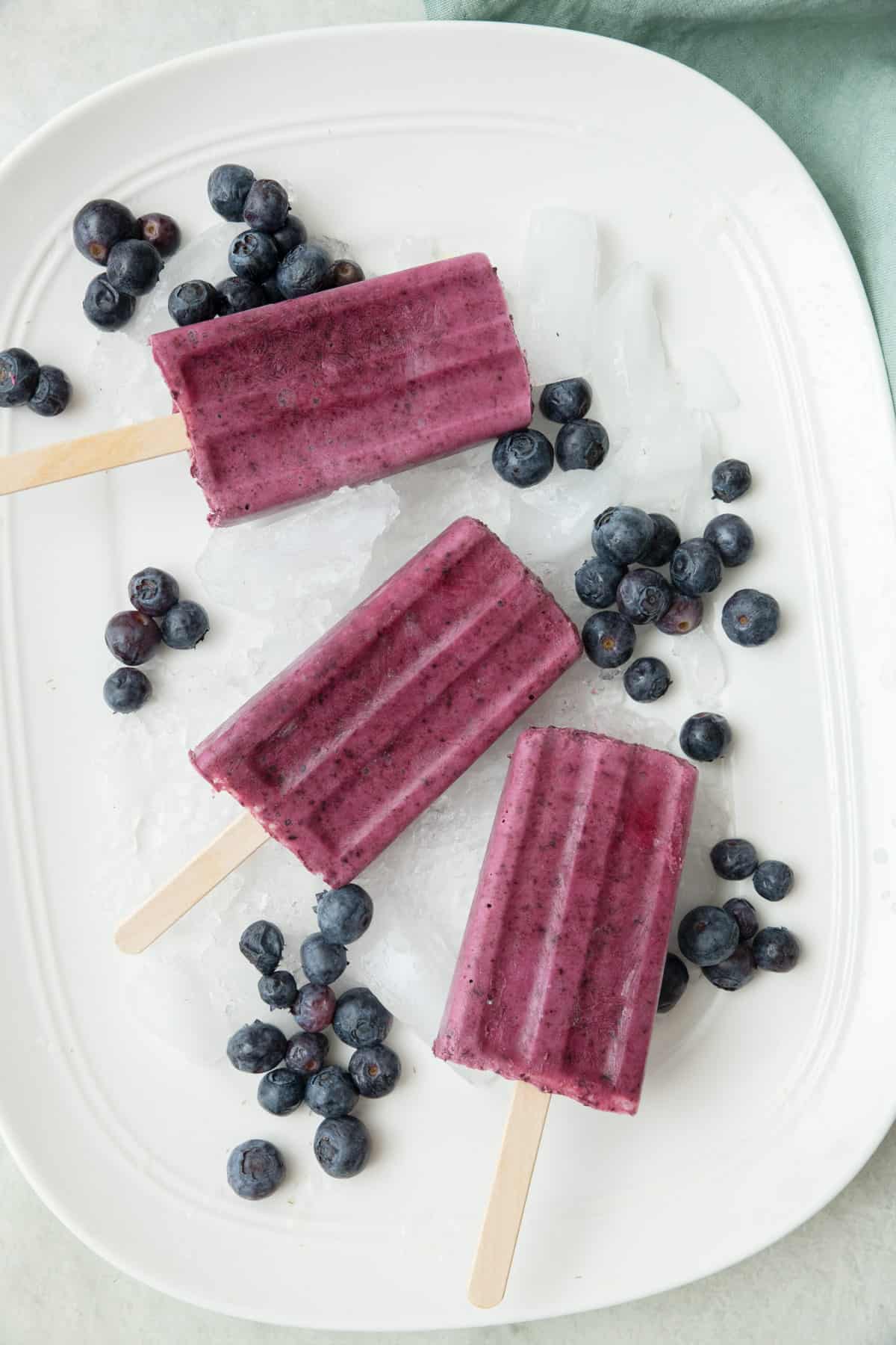 Frozen blueberry yogurt popsicles on a tray of ice with extra blueberries around.