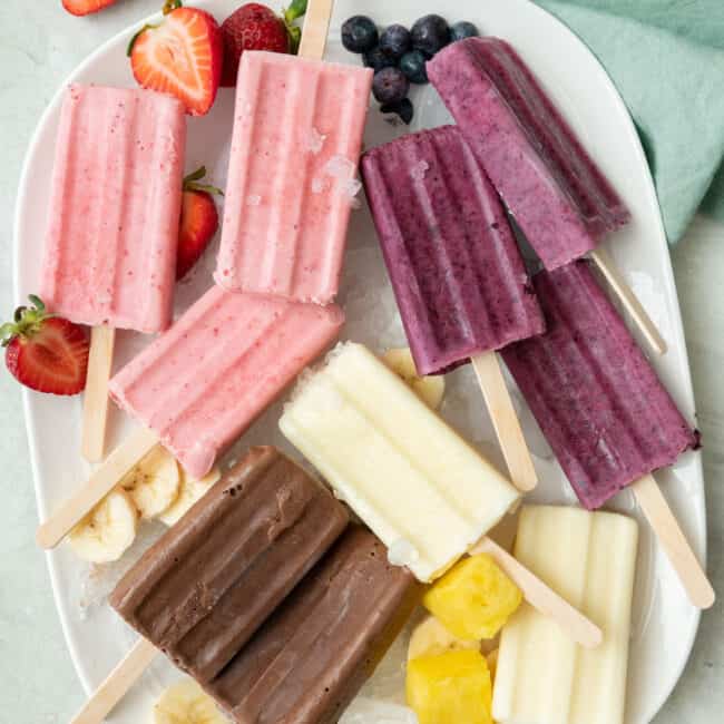 A variety of frozen yogurt popsicles on a large platter with extra main ingredients nearby: strawberry, blueberry, pineapple, and chocolate banana.