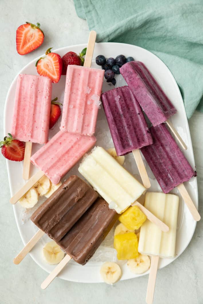 A variety of frozen yogurt popsicles on a large platter with extra main ingredients nearby: strawberry, blueberry, pineapple, and chocolate banana.