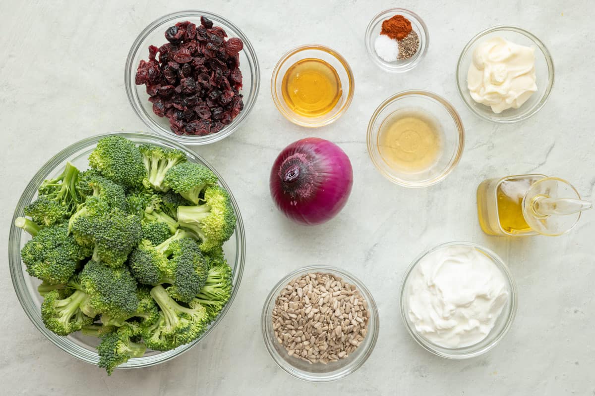 Ingredients for recipe before prepping in individual bowls: broccoli florets, raisins, vinegar, red onion, seeds, oil, honey, Greek yogurt, and mayonnaise.