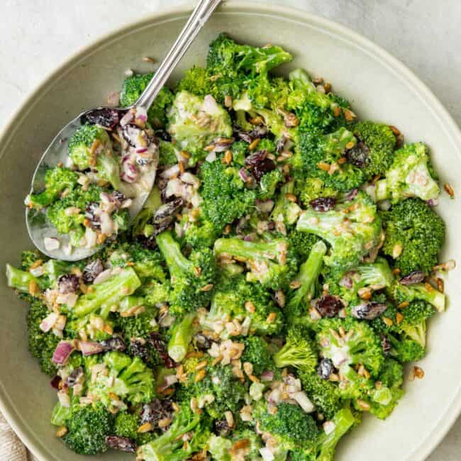 Broccoli salad in a large shallow bow with a serving spoon dipped in and an extra side of toasted sunflower seeds nearby.