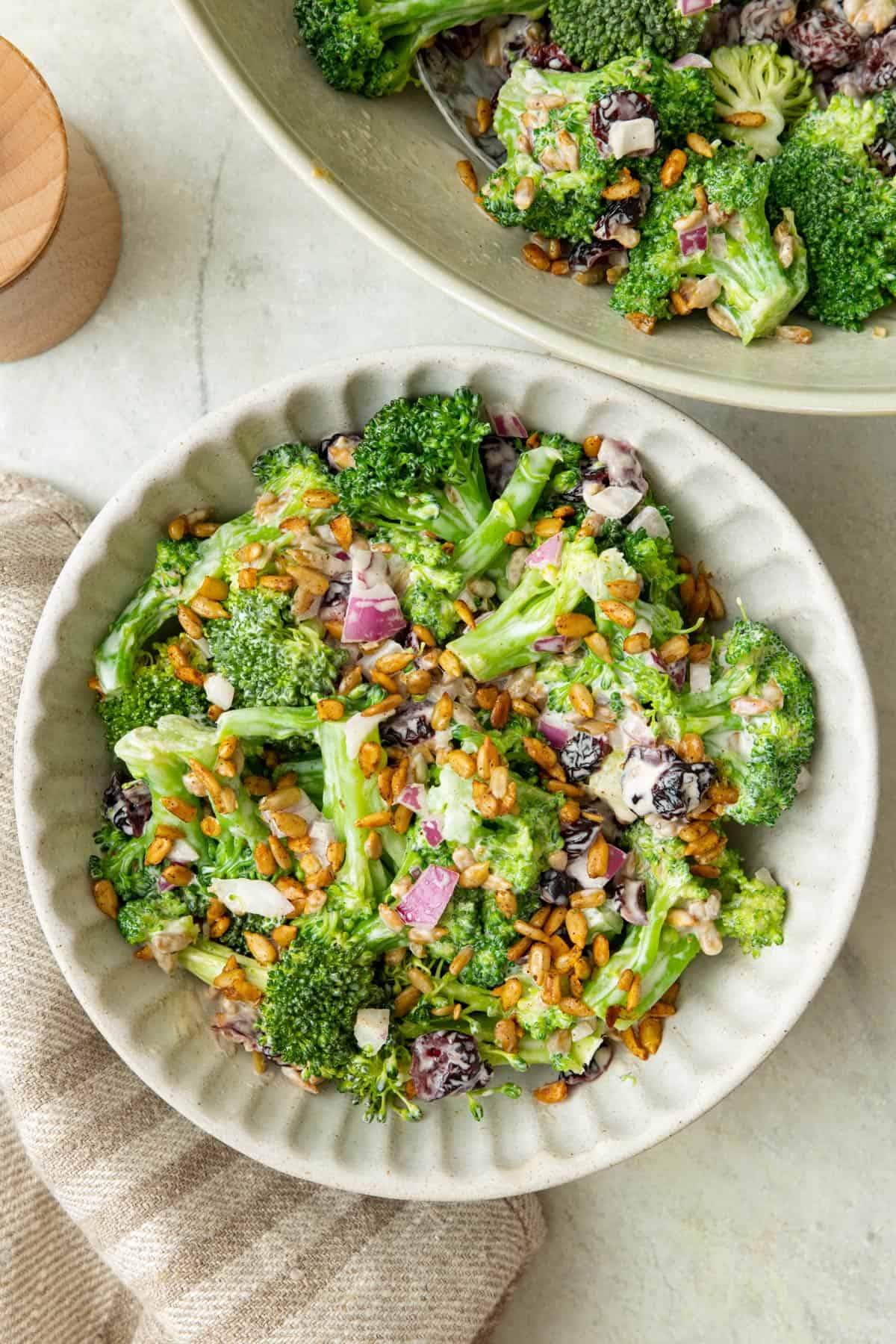Broccoli salad in a serving bowl next to large salad bowl with extra toasted nuts on top.