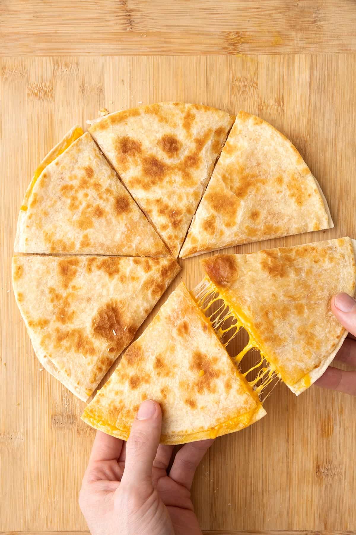 Cheese quesadilla on a cutting board cut in 6 pieces with hands pulling apart two slices to show a cheese pull.