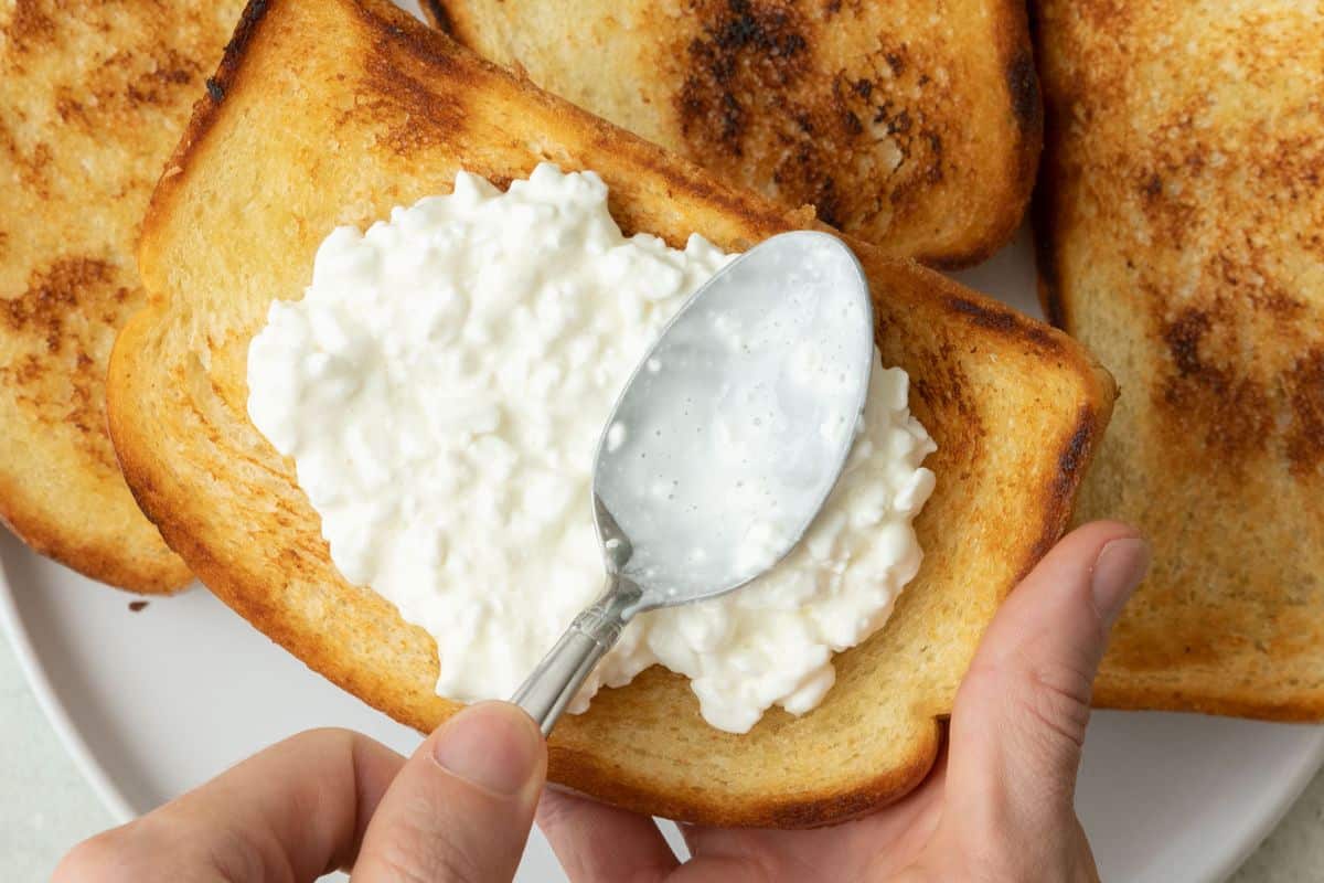 Cottage cheese being spread on to butter toast with a spoon.