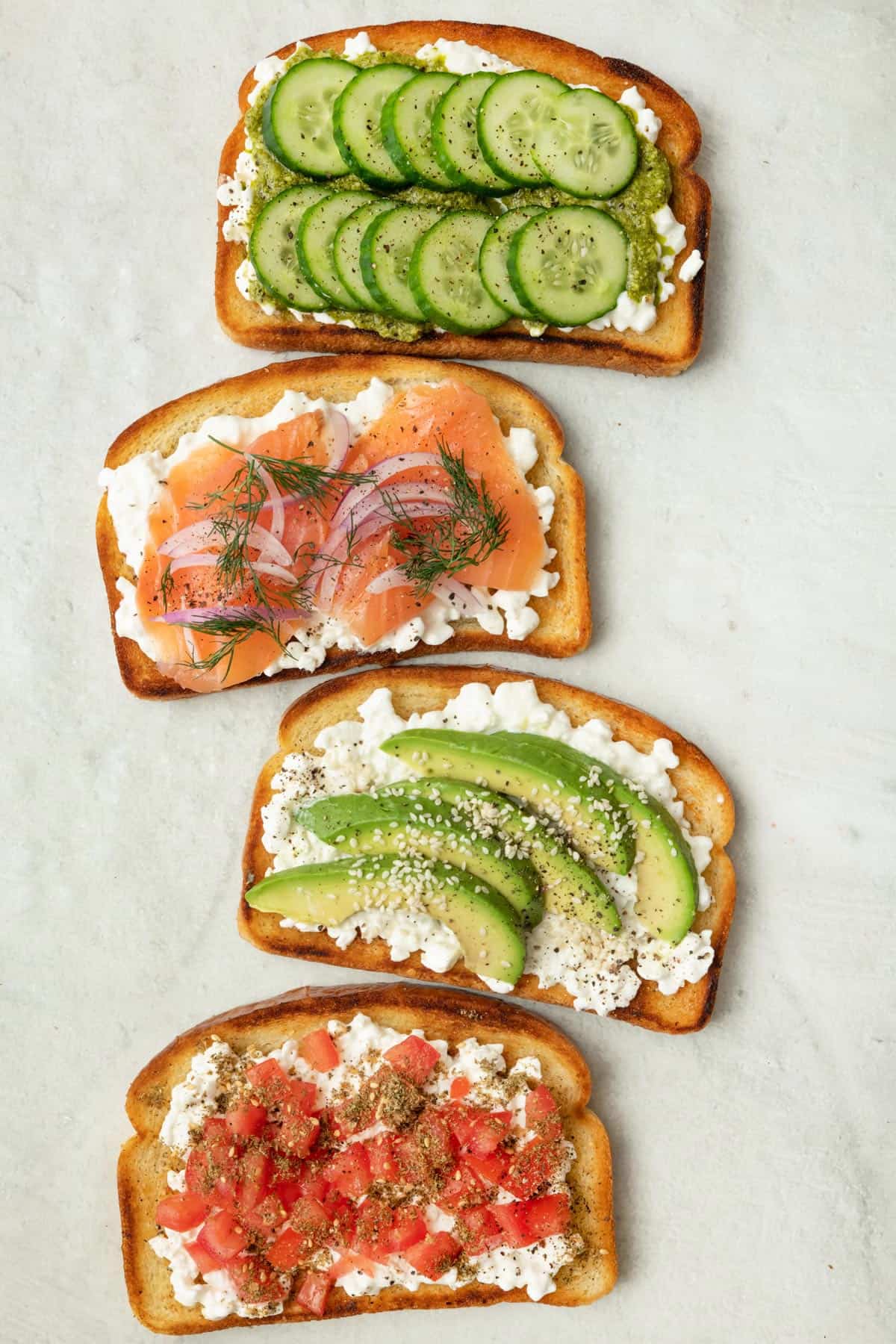 4 cottage cheese toasts with various toppings, from top to bottom: 1- sliced cucumbers and pesto, 2- smoked salmon, red onion, and fresh dill, 3- sliced avocado and sesame seeds, and 4- diced tomatoes and za'atar.