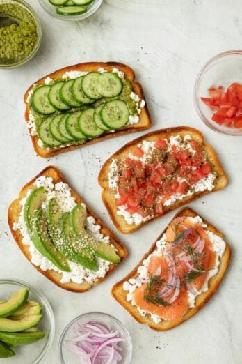 4 cottage cheese toasts with various toppings, from top to bottom: 1- sliced cucumbers and pesto, 2- smoked salmon, red onion, and fresh dill, 3- sliced avocado and sesame seeds, and 4- diced tomatoes and za'atar. Extra toppings in small bowls nearby.