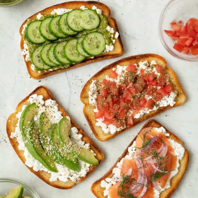 4 cottage cheese toasts with various toppings, from top to bottom: 1- sliced cucumbers and pesto, 2- smoked salmon, red onion, and fresh dill, 3- sliced avocado and sesame seeds, and 4- diced tomatoes and za'atar. Extra toppings in small bowls nearby.