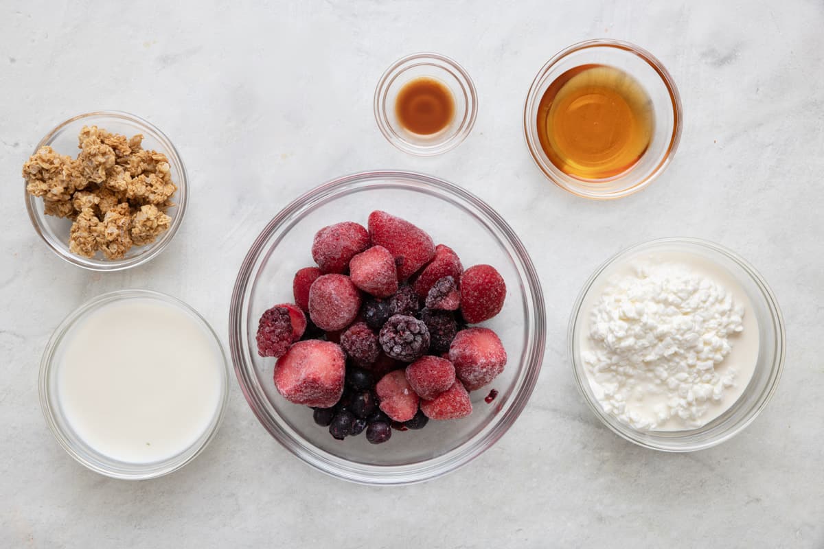 Ingredients for recipe in individual bowls: granola, milk, frozen mixed berries, vanilla extract, maple syrups, and cottage cheese.