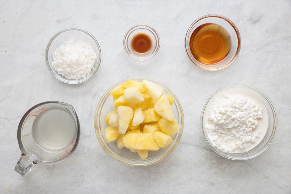 Ingredients for recipe in individual bowls: shredded coconut, coconut water, vanilla extract, frozen pineapple chunks, maple syrups, and cottage cheese.