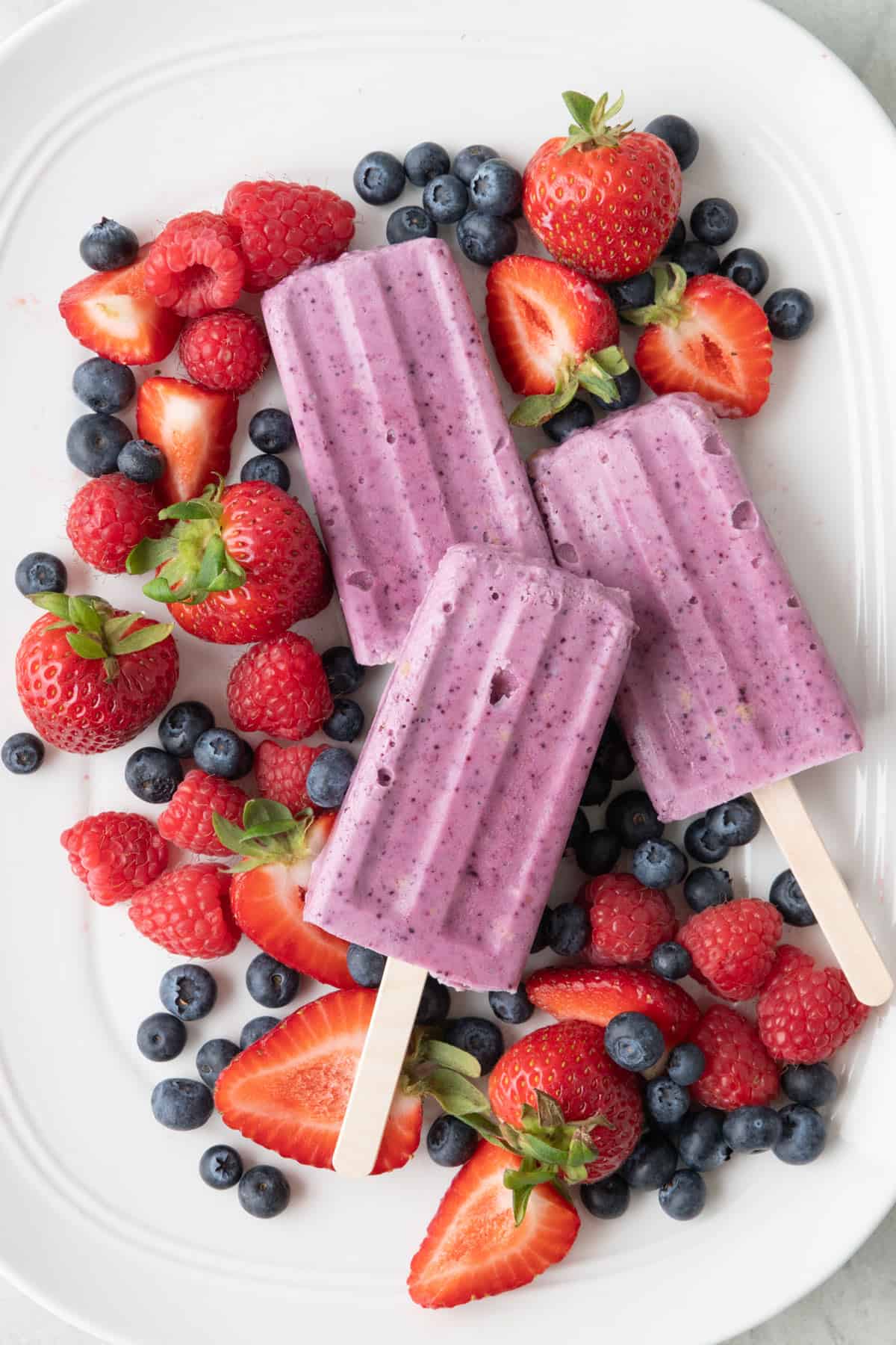 Frozen mixed berry cheesecake pops on a large platter with fresh mixed berries around.